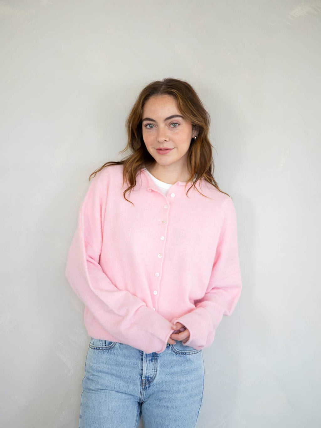 A female model wearing a light pink button up cardigan with white buttons, paired with light denim jeans in front of a gray studio background. 