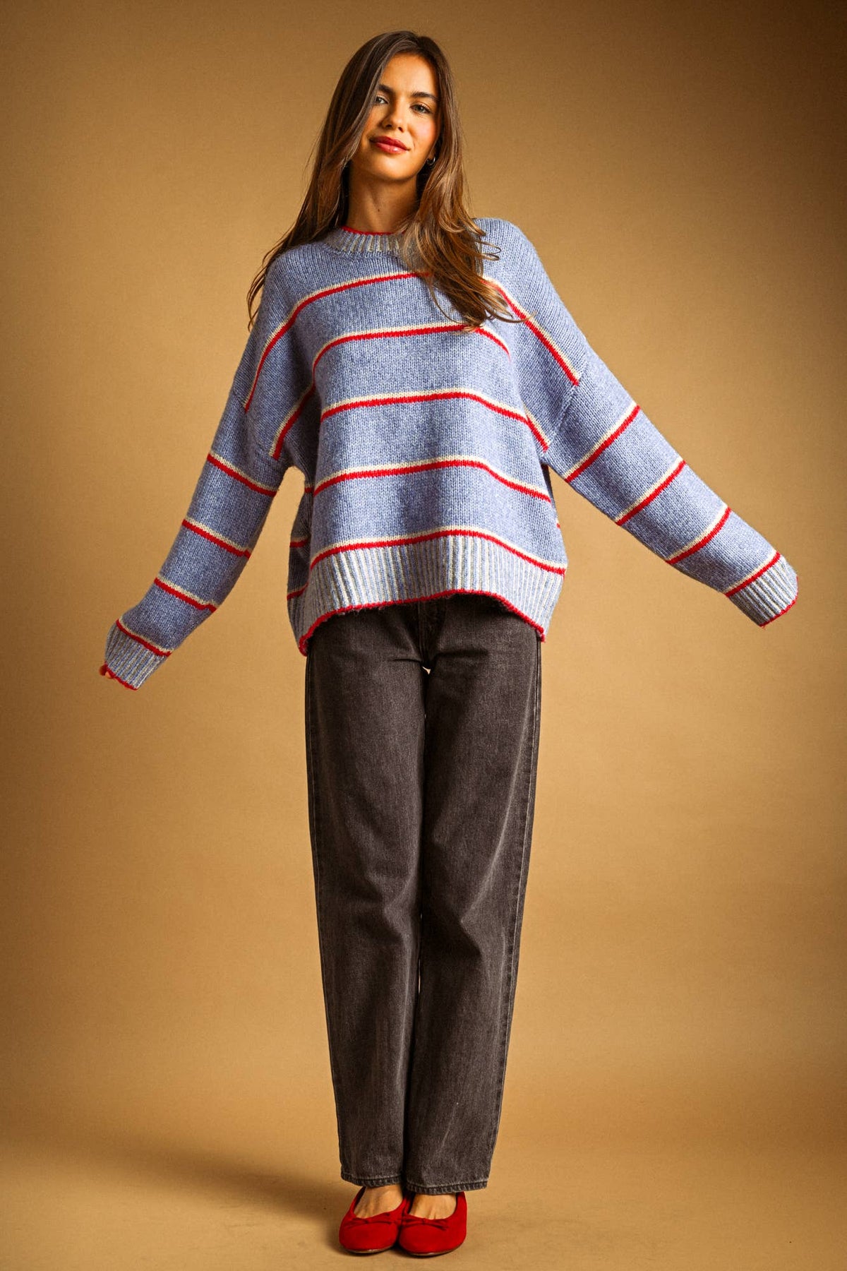A woman modeling a light blue knit sweater with red and white horizontal stripes, paired with dark gray wide-leg pants and red shoes, against a solid tan studio background.