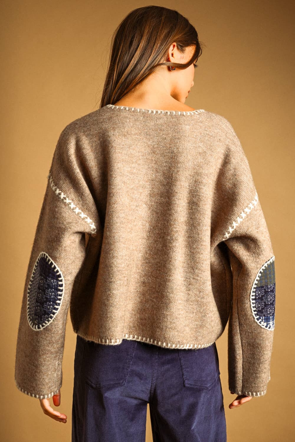 A woman viewed from behind wearing a cropped, heathered tan knit sweater with white decorative trim and blue patchwork elbow patches, paired with navy pants, against a solid tan background.