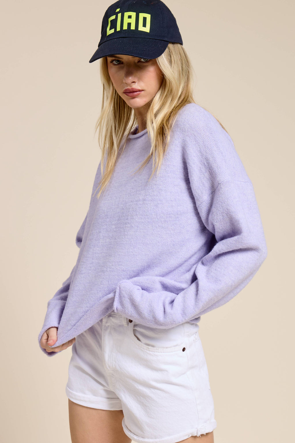 Woman wearing a lilac sweater and white shorts with a black cap on a beige background