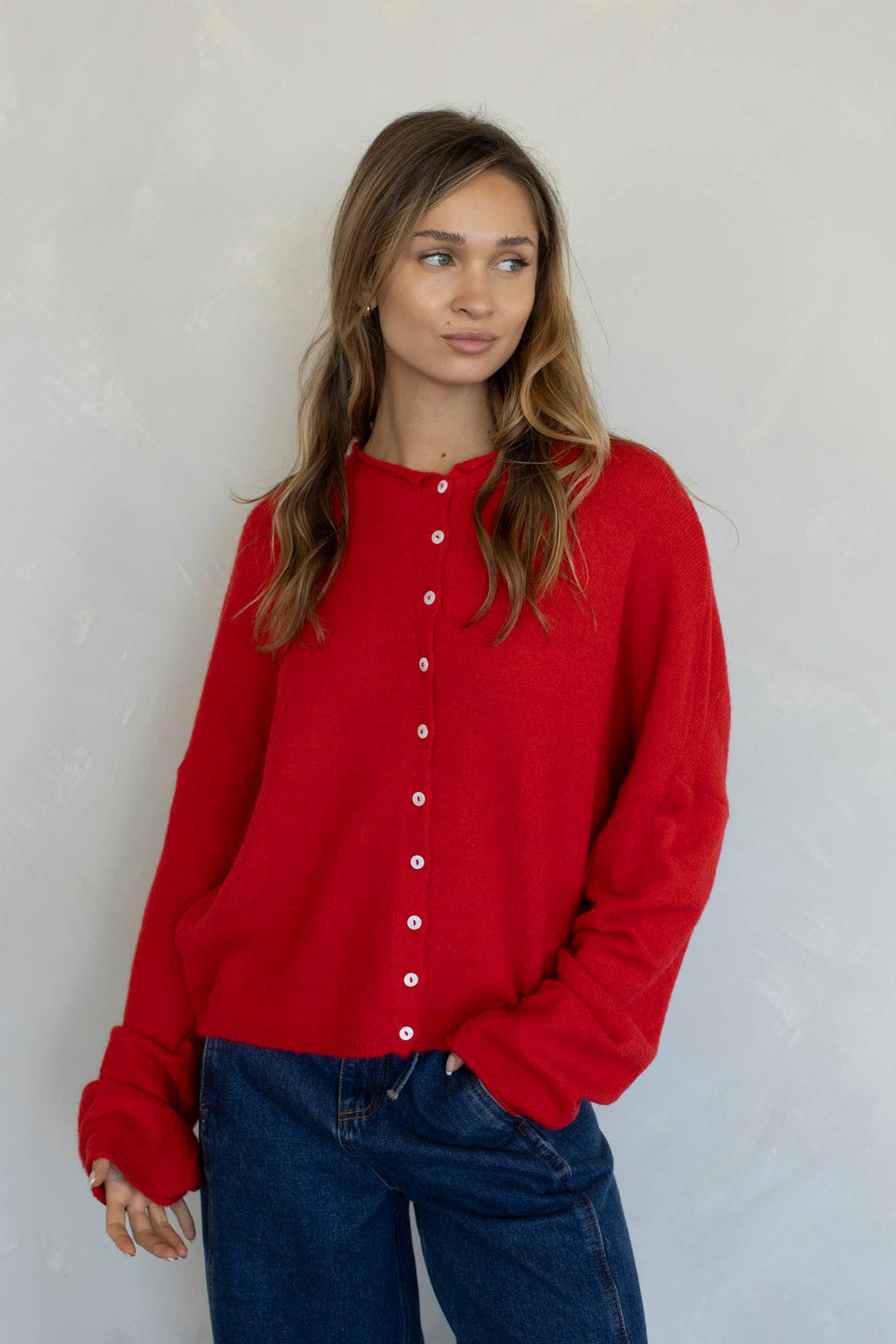 A female model wearing a bright red button up cardigan with white buttons, paired with dark denim jeans in front of a gray studio background. 