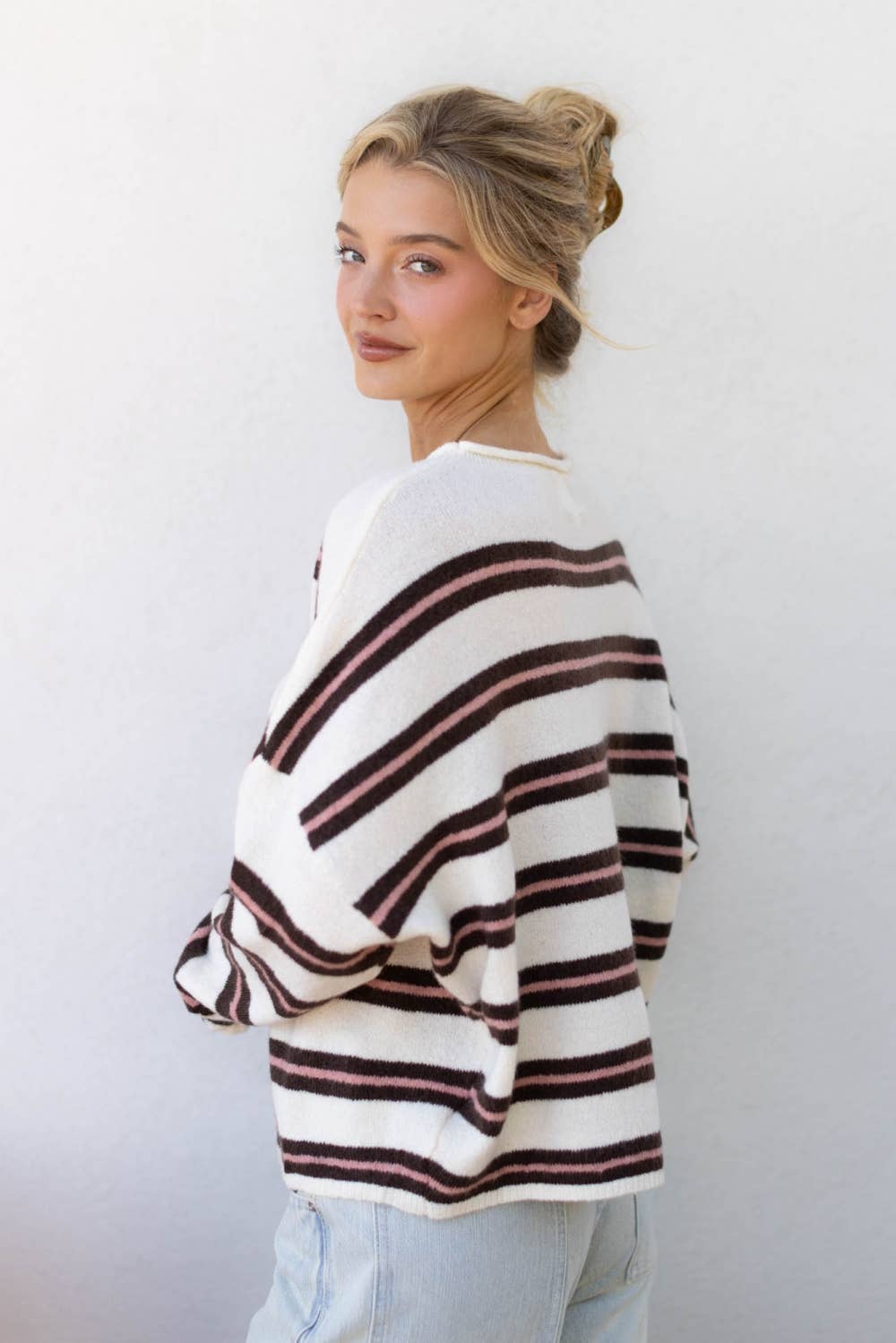 A woman wearing a white knit sweater with horizontal dark brown and dusty rose stripes, paired with light blue jeans, standing against a plain white wall.