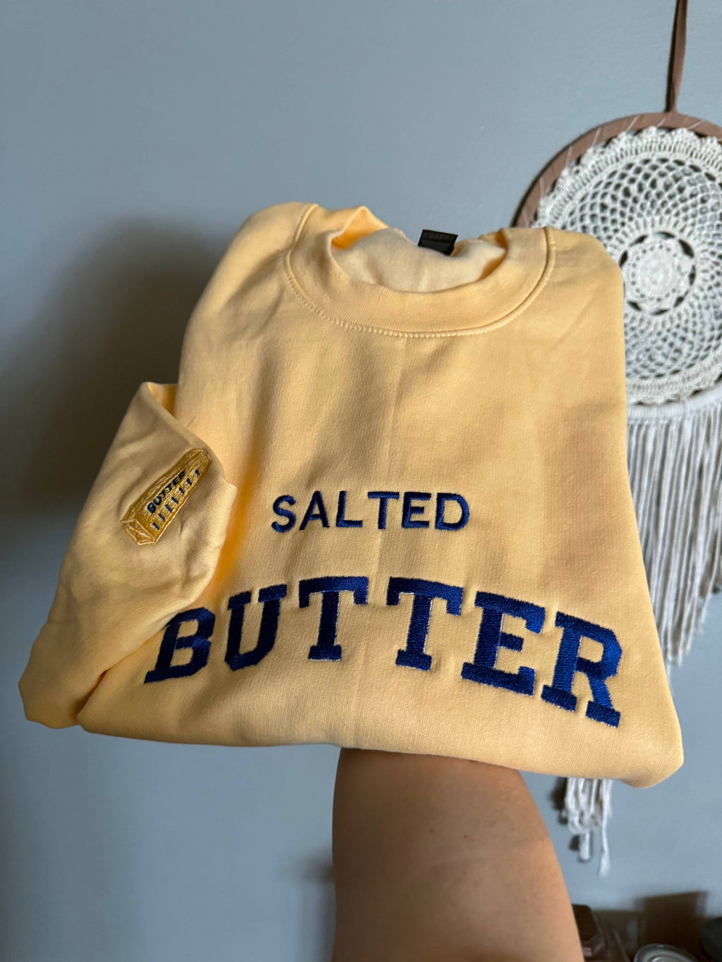 Yellow sweatshirt with 'SALTED BUTTER' in blue text held by a person against a gray wall.