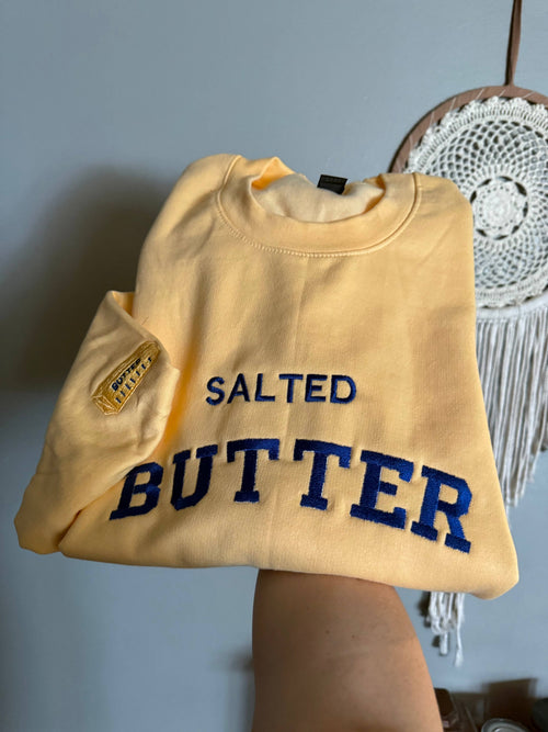 Yellow sweatshirt with 'SALTED BUTTER' in blue text held by a person against a gray wall.