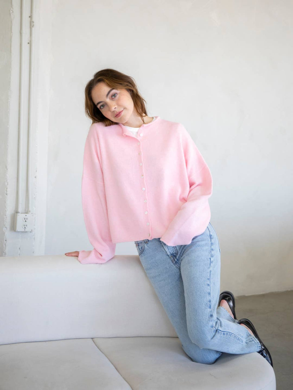 A female model kneeling on a couch, wearing a light pink button up cardigan with white buttons, paired with light denim jeans in front of a gray studio background. 