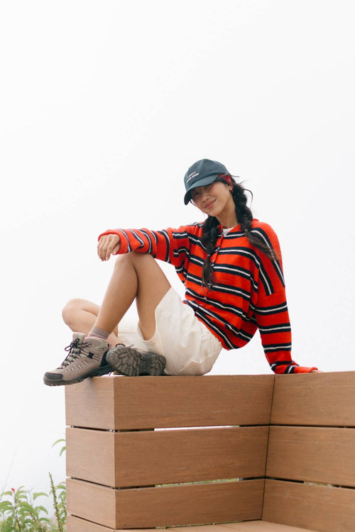 A woman sits on a wooden bench wearing a red, black, and white striped long-sleeve top, white shorts, hiking shoes, and a dark baseball cap against a bright white sky.