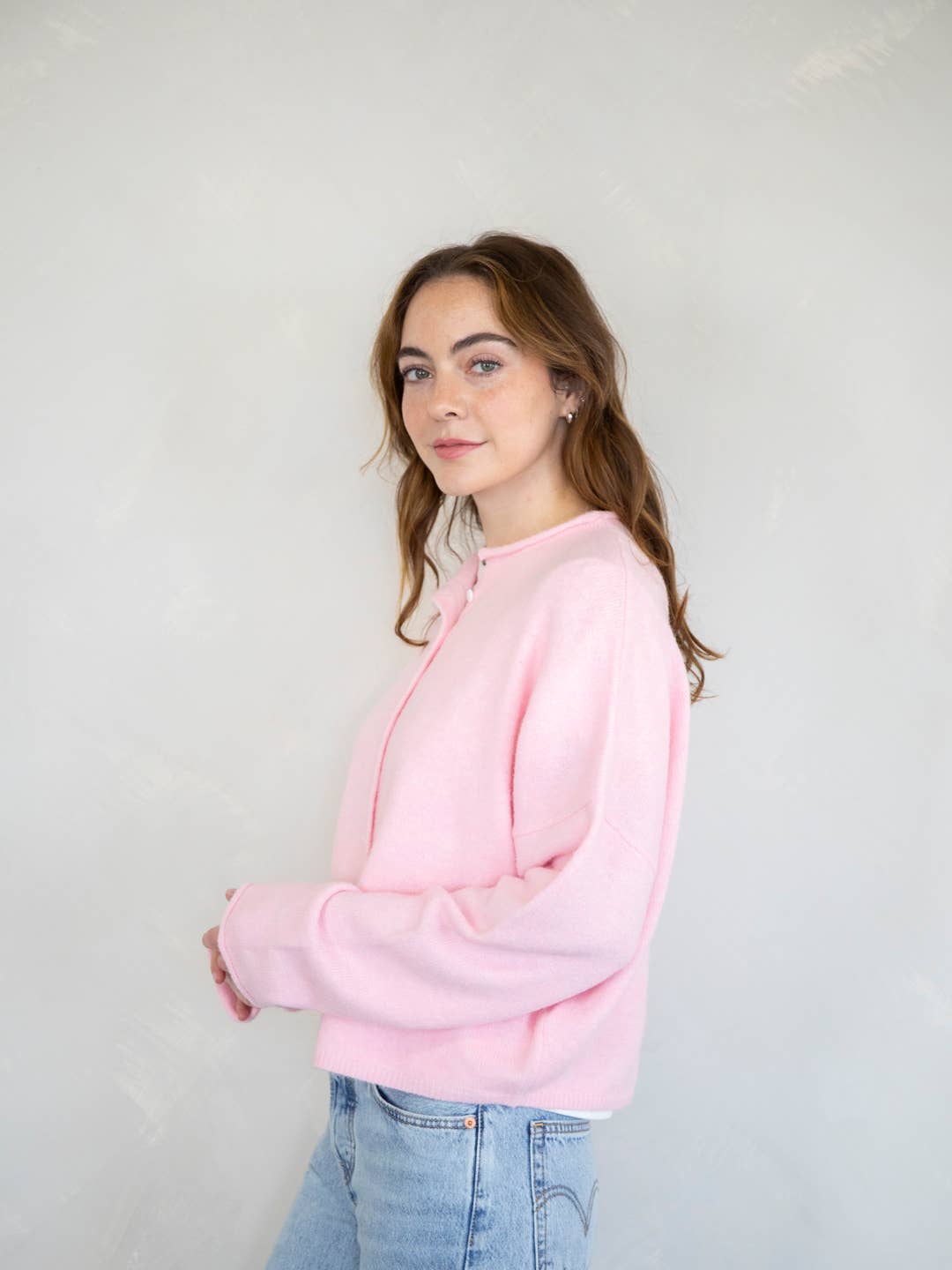 A female model wearing a light pink button up cardigan with white buttons, paired with light denim jeans in front of a gray studio background. 