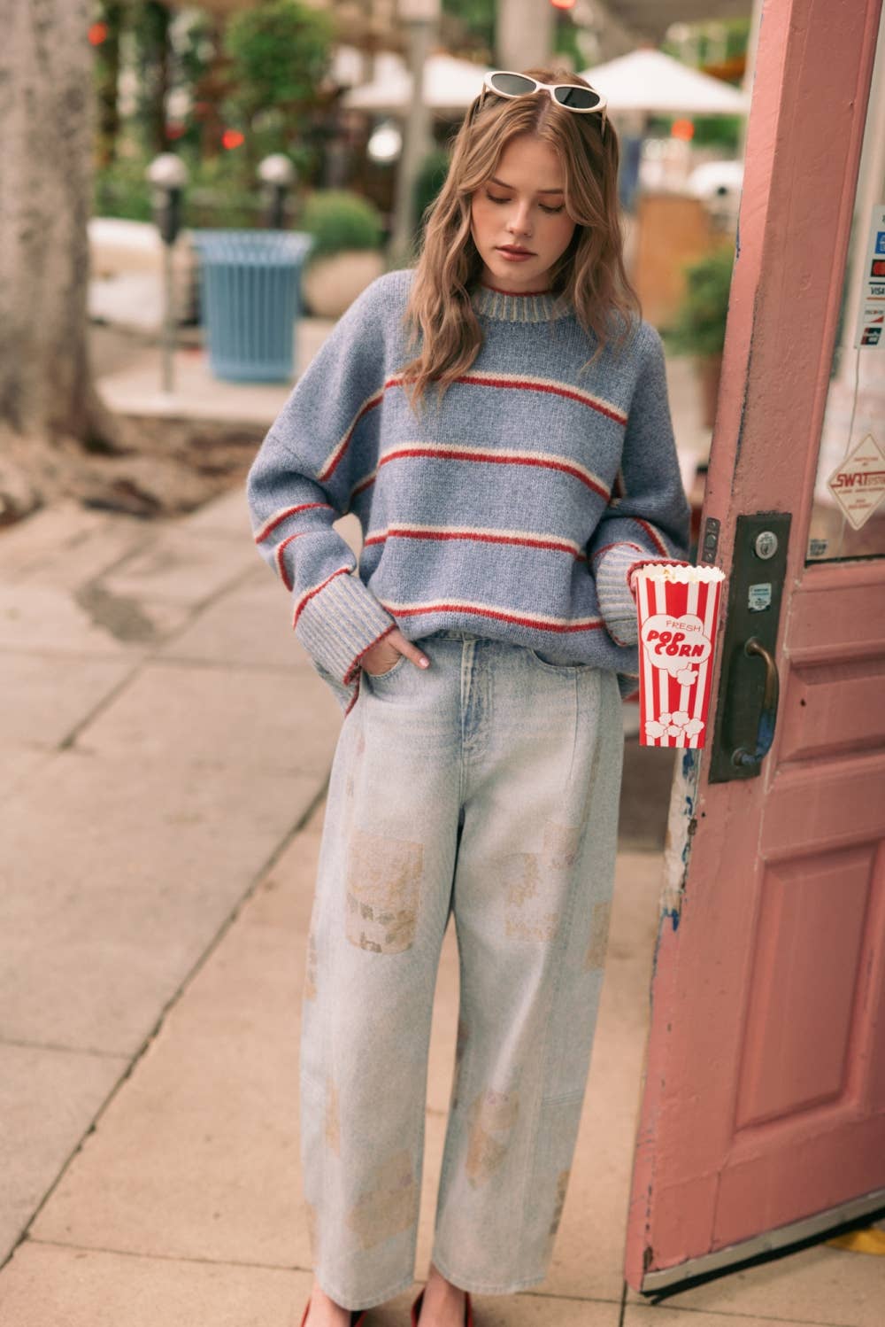 A woman stands on a sidewalk next to a pink door, wearing a blue knit sweater with red and white horizontal stripes, light-wash distressed jeans, and sunglasses on her head. She is holding a red and white popcorn bucket.