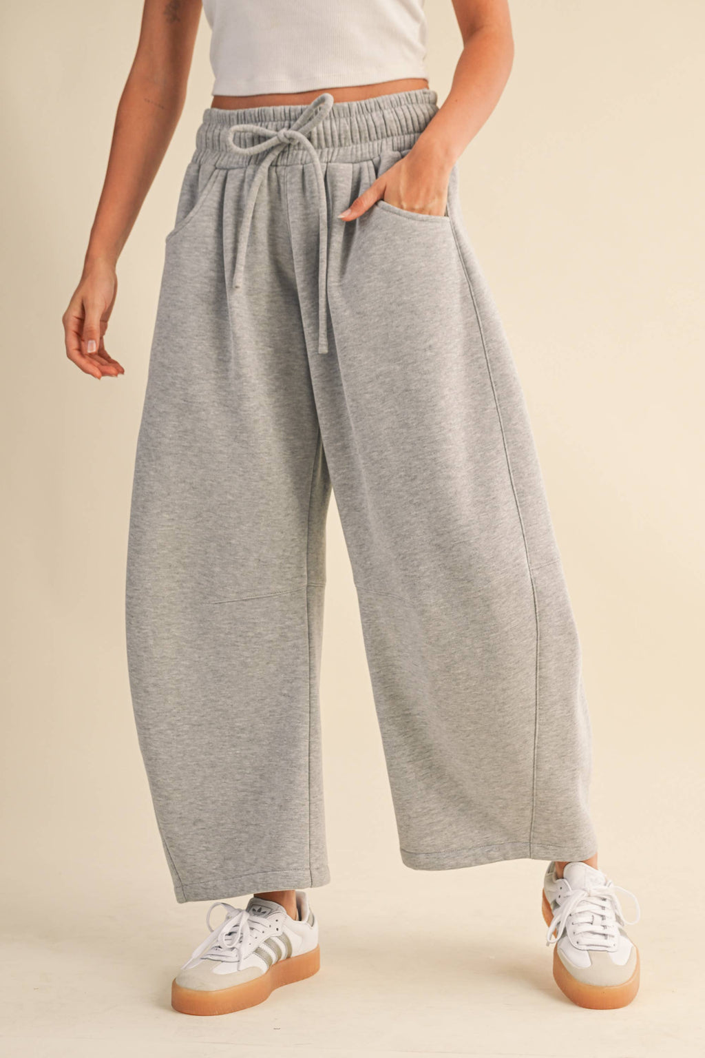 A person wearing light heather gray wide-leg barrel sweatpants with an elastic drawstring waist and pockets, paired with white sneakers, against a plain beige studio background.