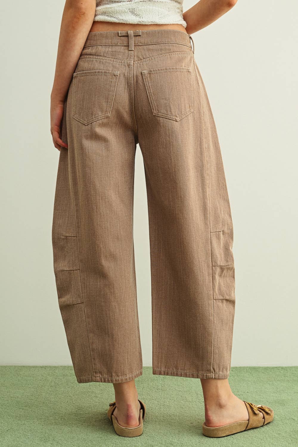 A person wearing wide-leg barrel cropped pants in a warm taupe-brown color, paired with tan double-strap sandals and a light-colored knit top, standing against a plain light wall with a green floor while facing backwards. 