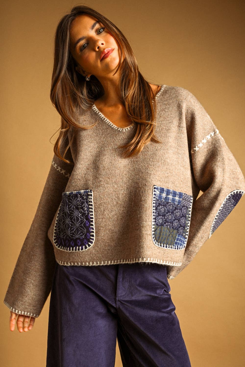 A woman wearing a beige knit sweater with blue patchwork pockets and dark purple pants, posed against a solid warm brown background.