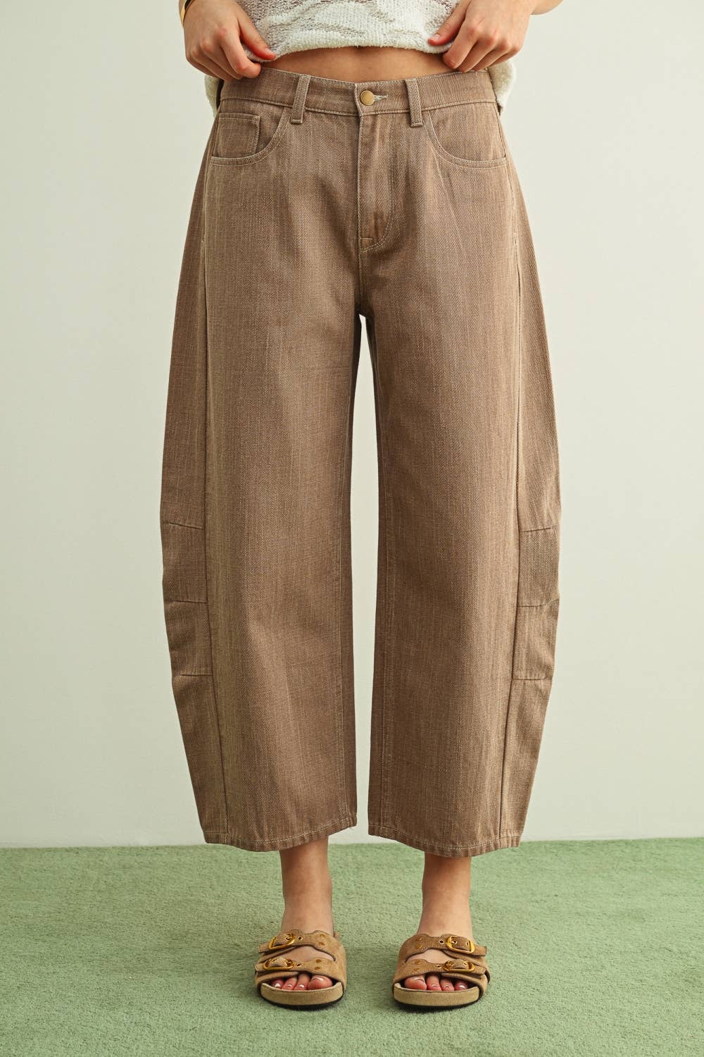 A person wearing wide-leg barrel cropped pants in a warm taupe-brown color, paired with tan double-strap sandals and a light-colored knit top, standing against a plain light wall with a green floor.