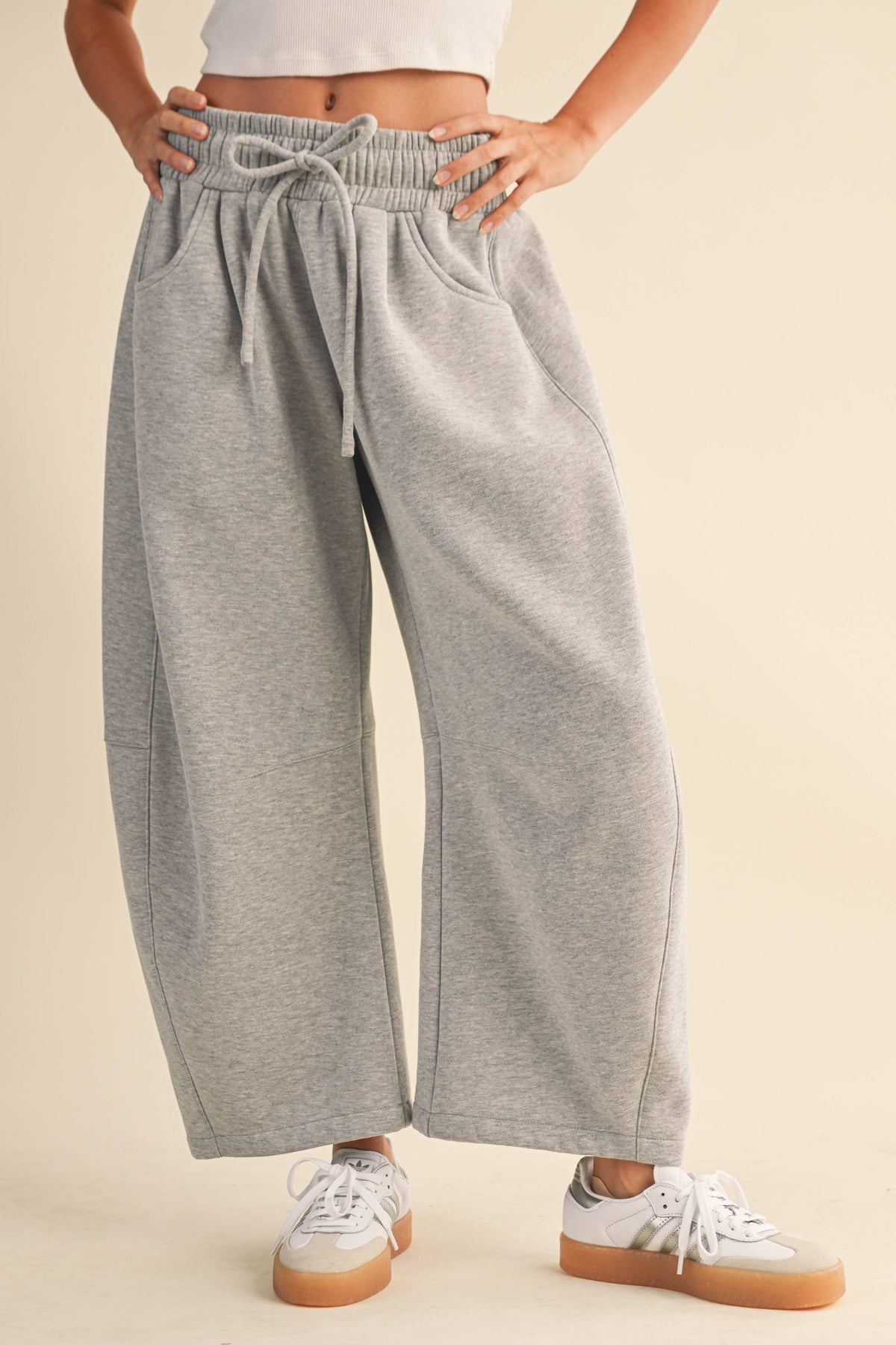 A person wearing light heather gray wide-leg barrel sweatpants with an elastic drawstring waist and side pockets, paired with white sneakers, against a plain beige studio background.