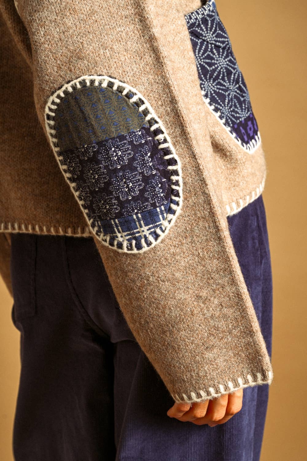 Close-up of a beige wool sweater with dark blue patchwork elbow patches and white contrast stitching, worn by a model with dark navy trousers against a warm tan background.