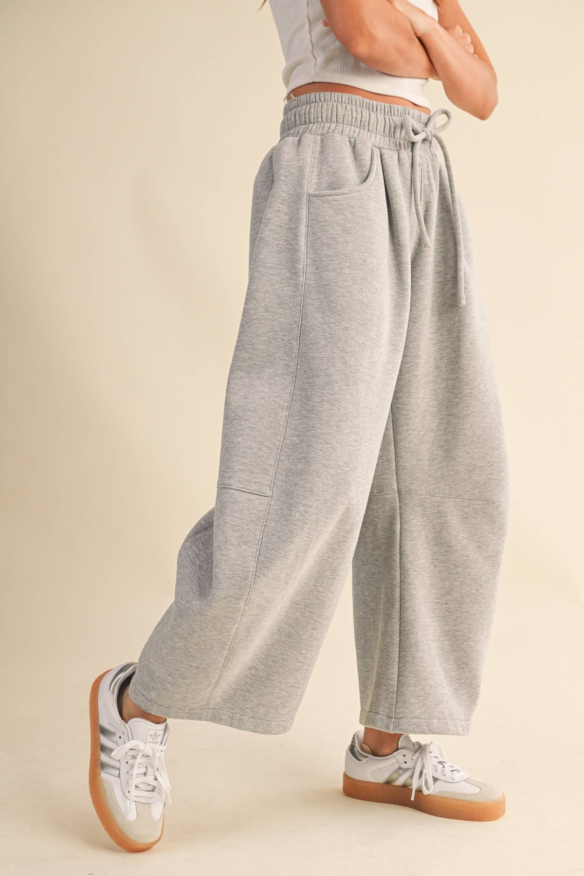 A person wearing light heather gray wide-leg barrel sweatpants with an elastic drawstring waist and side pockets, paired with white sneakers, shown from the waist down against a plain beige studio background.