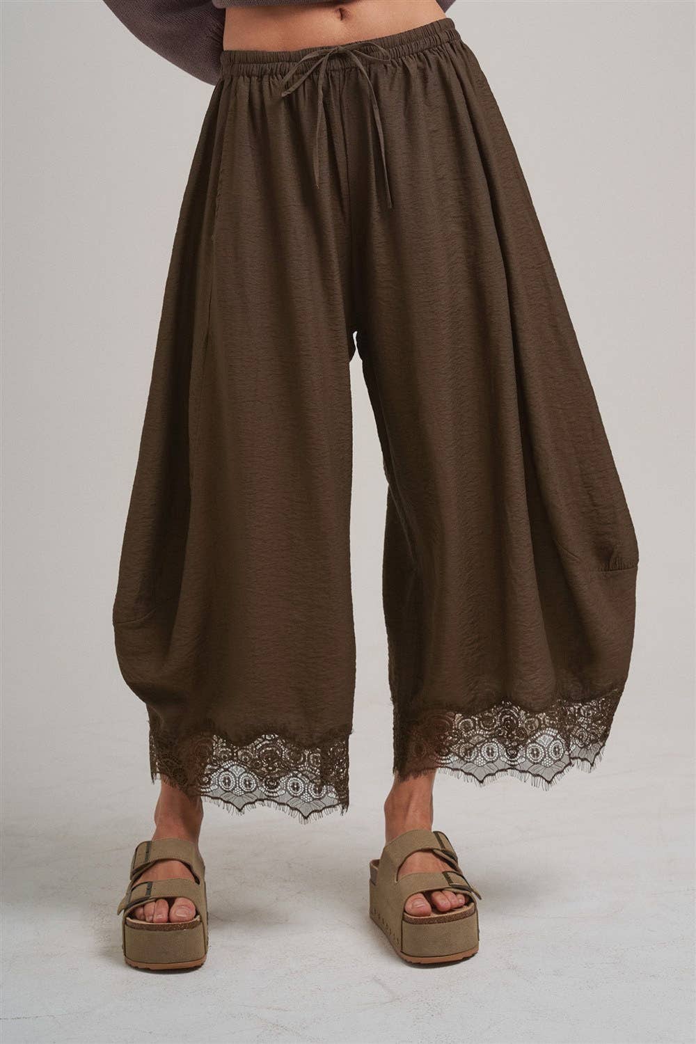 Brown wide-leg pants with lace trim worn by a person who is also wearing tan platform sandals on a plain background