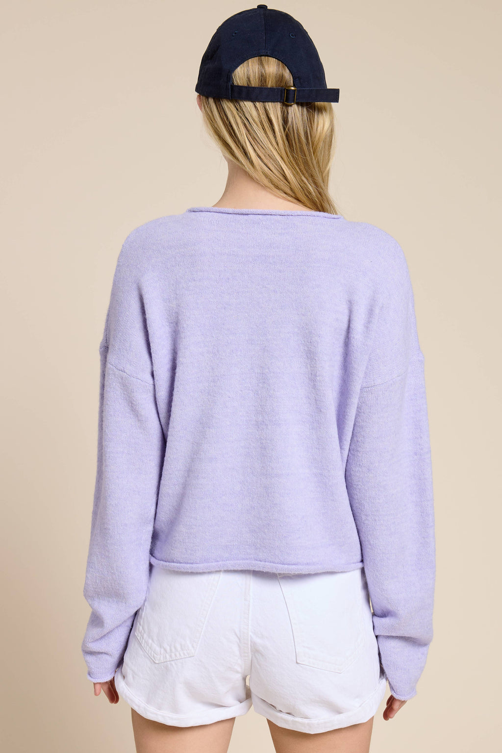 Person facing backwards wearing a light purple sweater and white shorts with a navy cap on a beige background