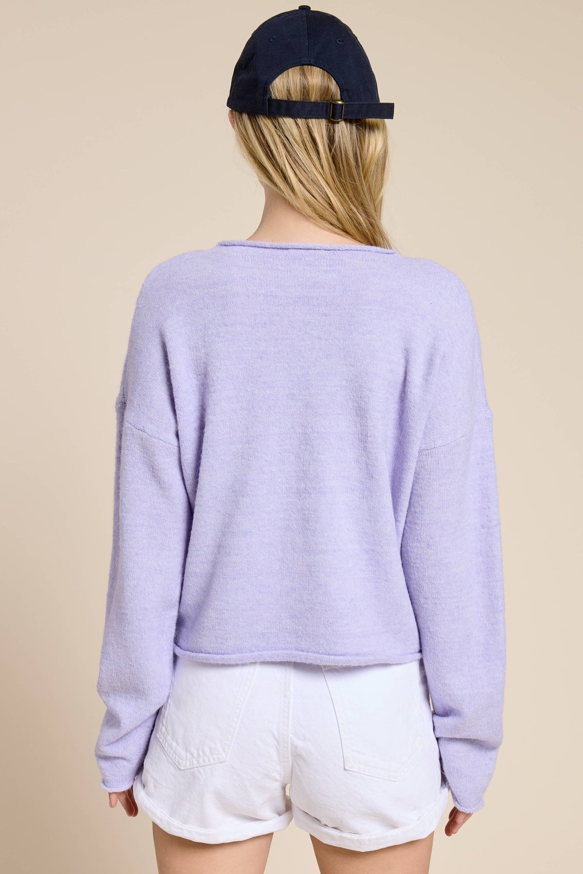 Person facing backwards wearing a light purple sweater and white shorts with a navy cap on a beige background