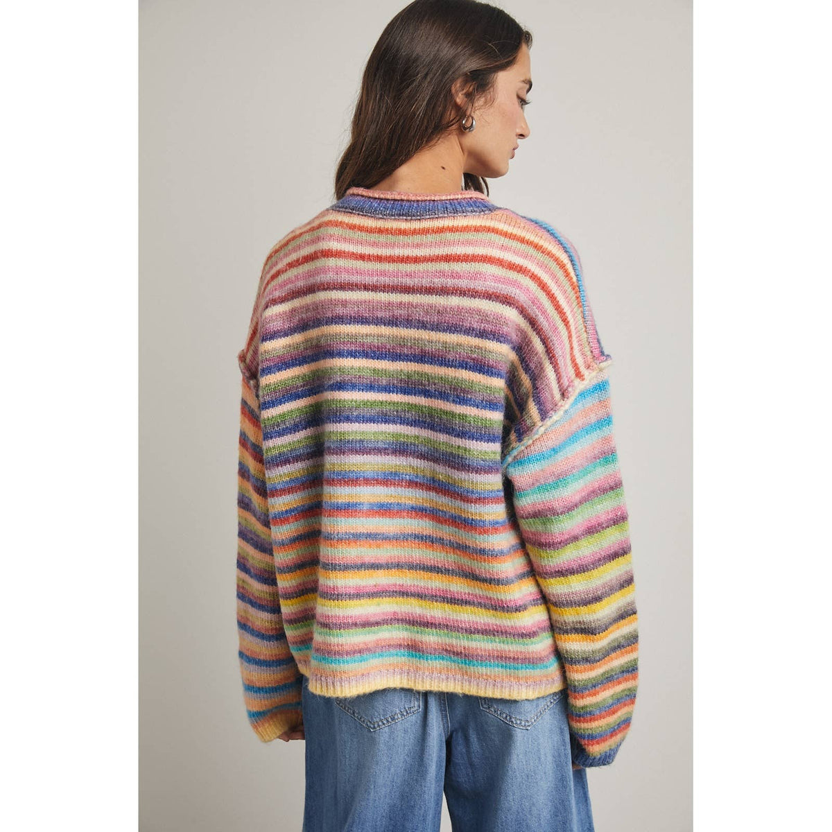 A woman facing backwards wearing a colorful, rainbow fuzzy horizontal striped cardigan sweater paired with blue jeans, against a plain light gray background.