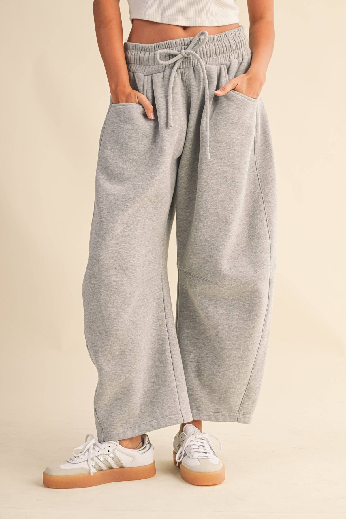 A person wearing light heather gray wide-leg barrel sweatpants with an elastic drawstring waist and side pockets, paired with white sneakers, against a plain beige studio background.