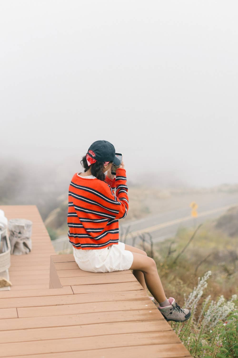 A woman sitting on a wooden boardwalk wearing a red, white, and black striped long-sleeve shirt, white shorts, and a black cap, looking out over a foggy mountain landscape.