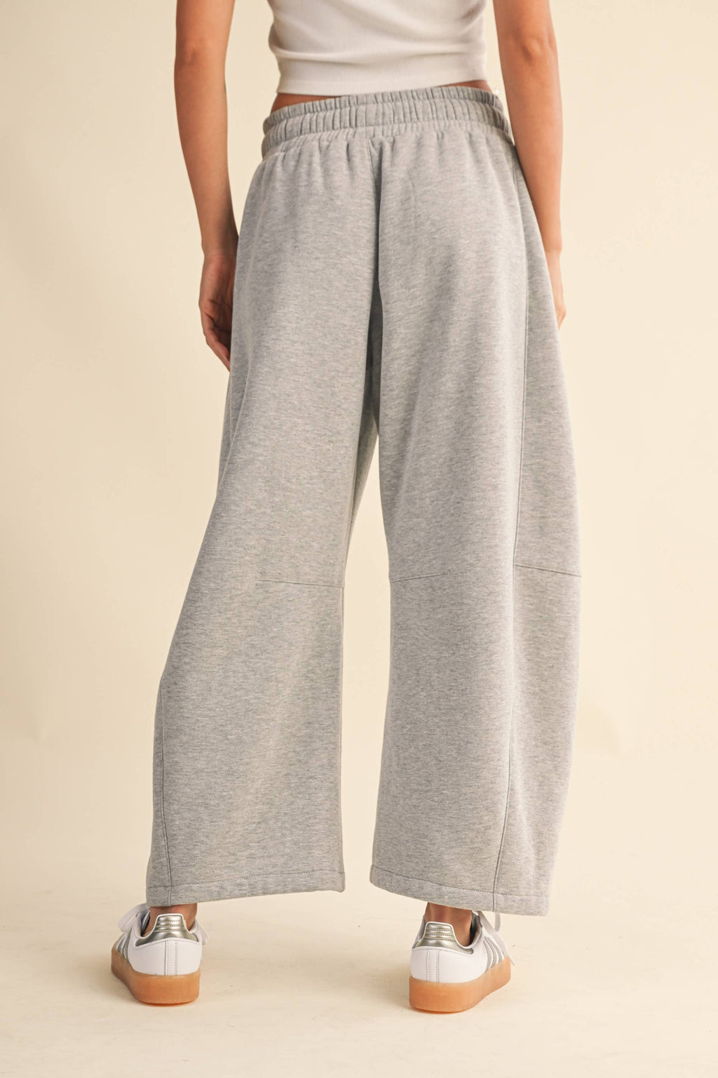 A person shown from the back wearing light heather gray wide-leg barrel sweatpants, a white top, and white sneakers with tan soles, against a plain beige studio background.