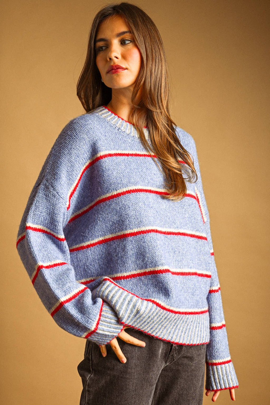 A woman wearing a light blue knit sweater with horizontal red and white stripes, paired with dark gray pants, set against a solid warm brown background.
