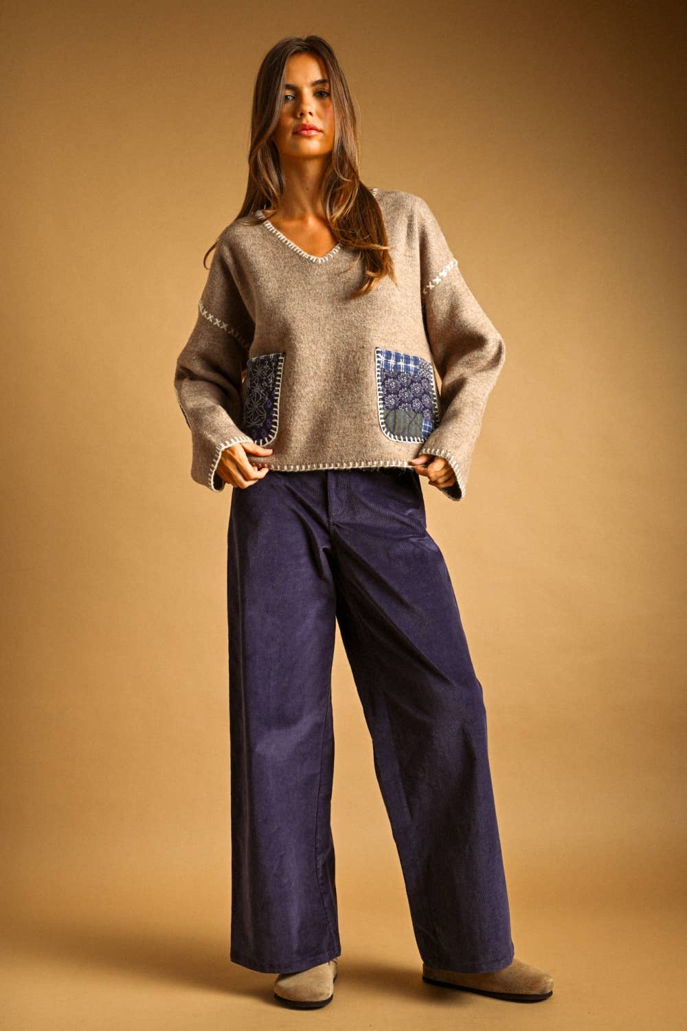 A woman modeling a beige knit sweater with blue patterned front pockets and wide-leg navy pants, standing against a warm tan studio backdrop.