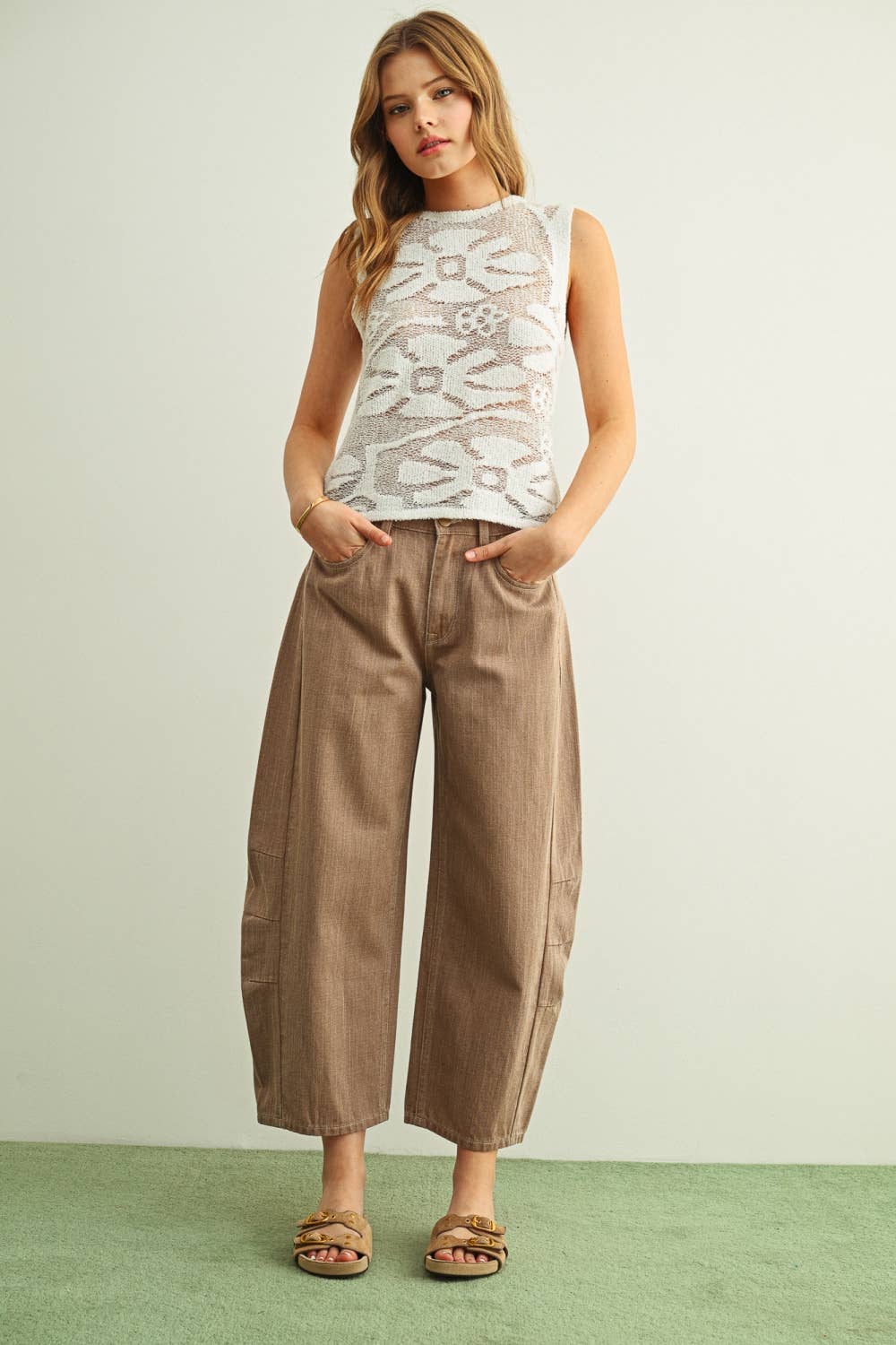 A person wearing wide-leg barrel cropped pants in a warm taupe-brown color, paired with tan double-strap sandals and a light-colored knit top, standing against a plain light wall with a green floor.