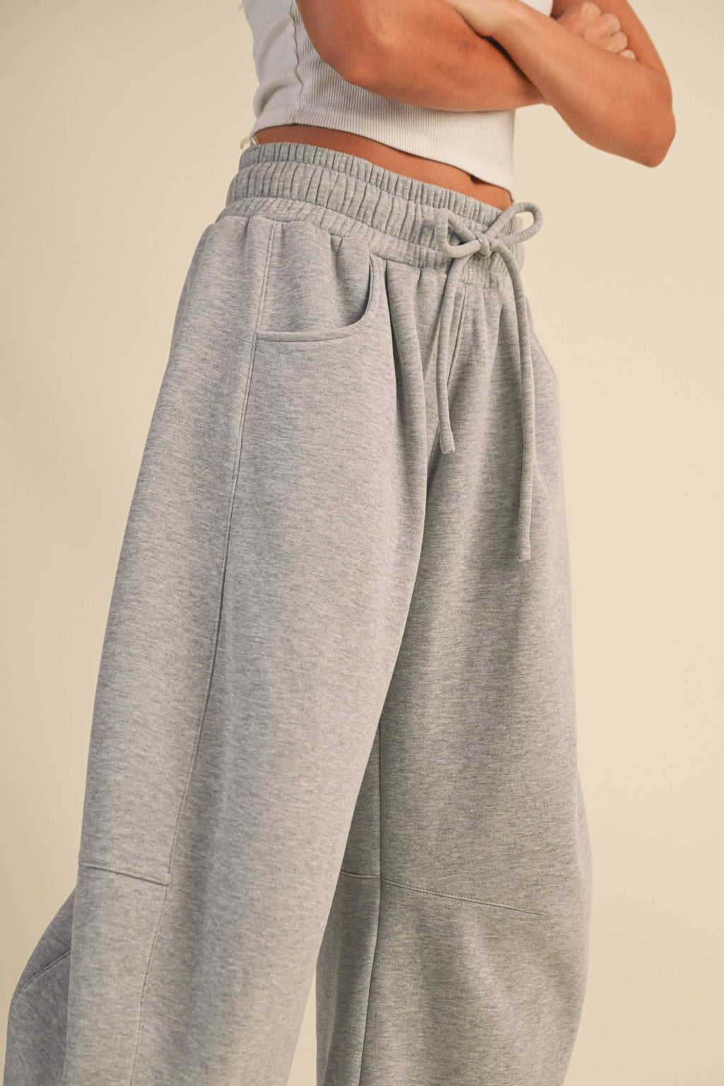 Close-up of a person wearing light heather gray drawstring sweatpants with a ribbed elastic waistband and side pockets, paired with a white cropped top against a plain beige studio background.