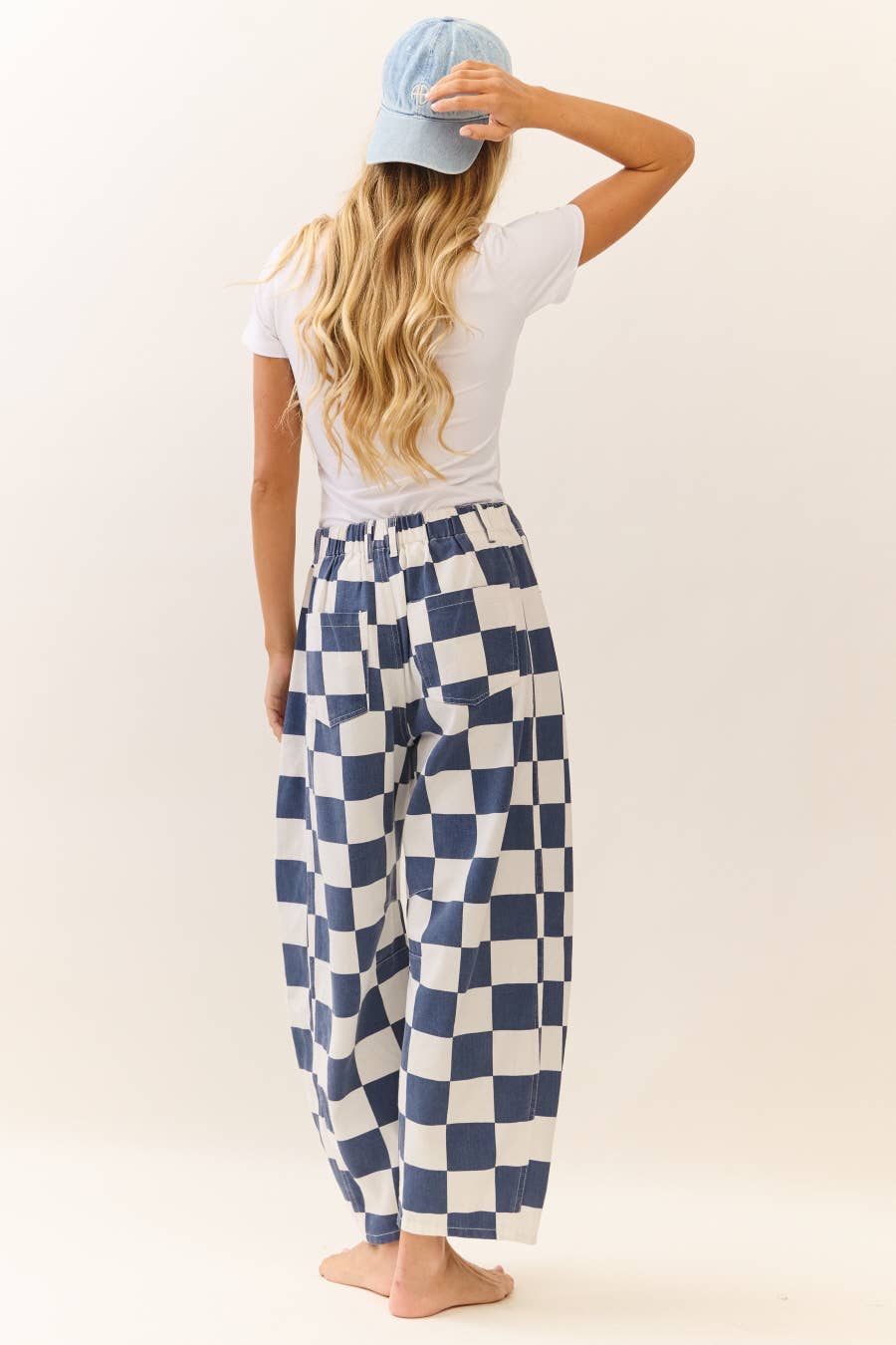 Woman facing backwards wearing blue and white checkered barrel pants with a white top on a plain background