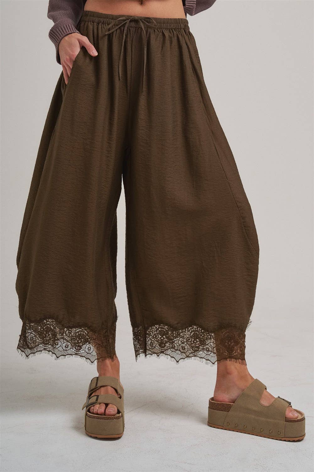 A person wearing loose, wide-leg brown harem-style pants with a lace hem, paired with brown platform sandals against a plain light gray studio background.