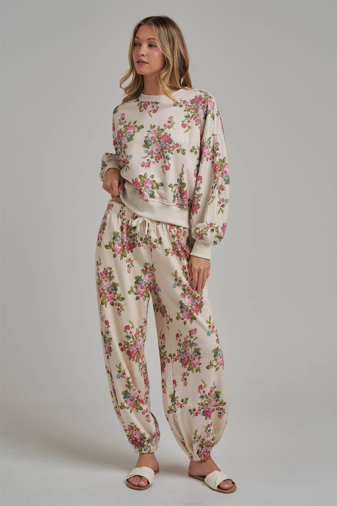Floral patterned wide leg jogger sweat pants and matching pullover sweatshirt worn by a female model with white sandals on a plain background