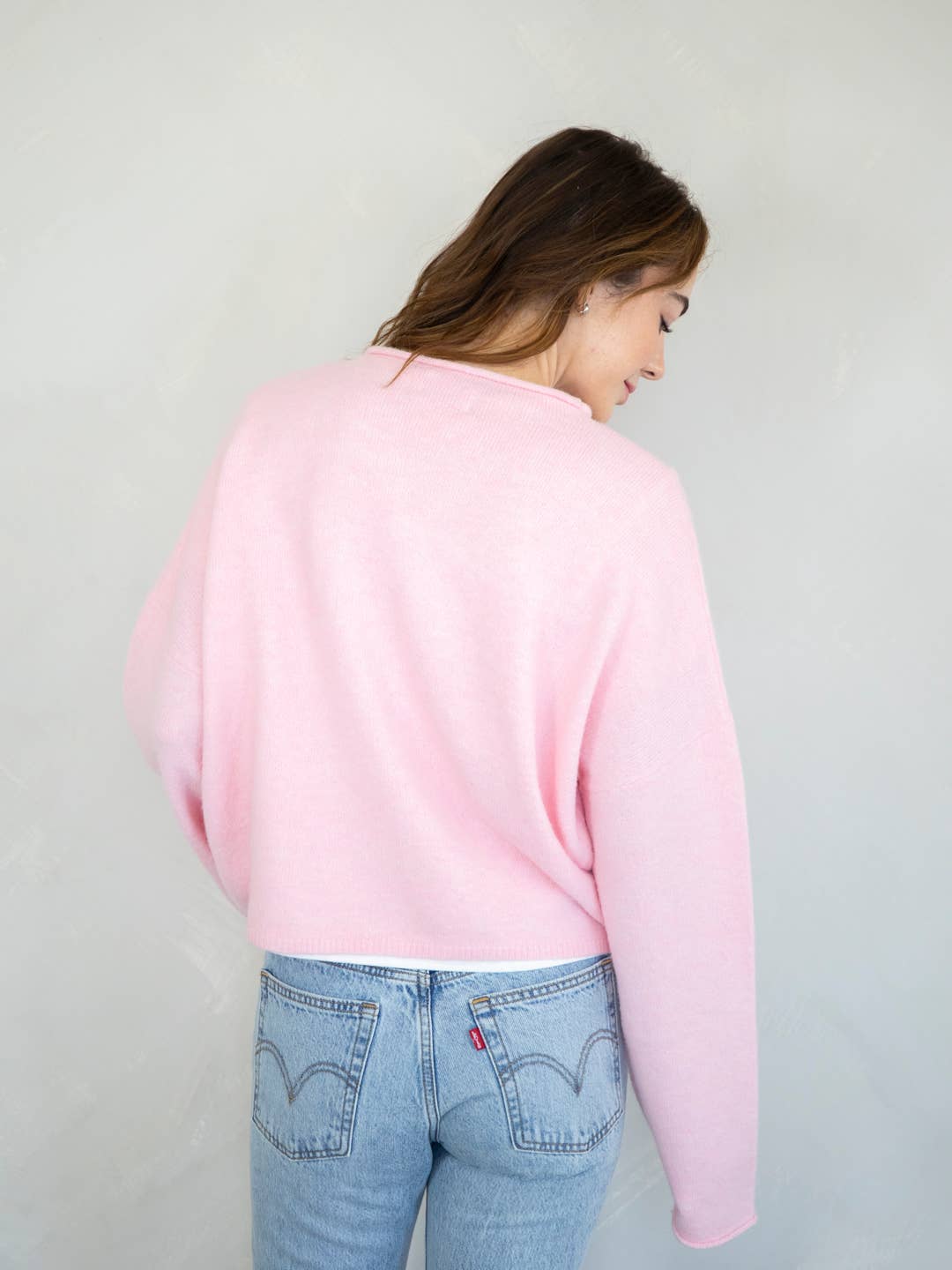 A female model facing backwards, wearing a light pink cardigan paired with dark denim jeans in front of a gray studio background. 