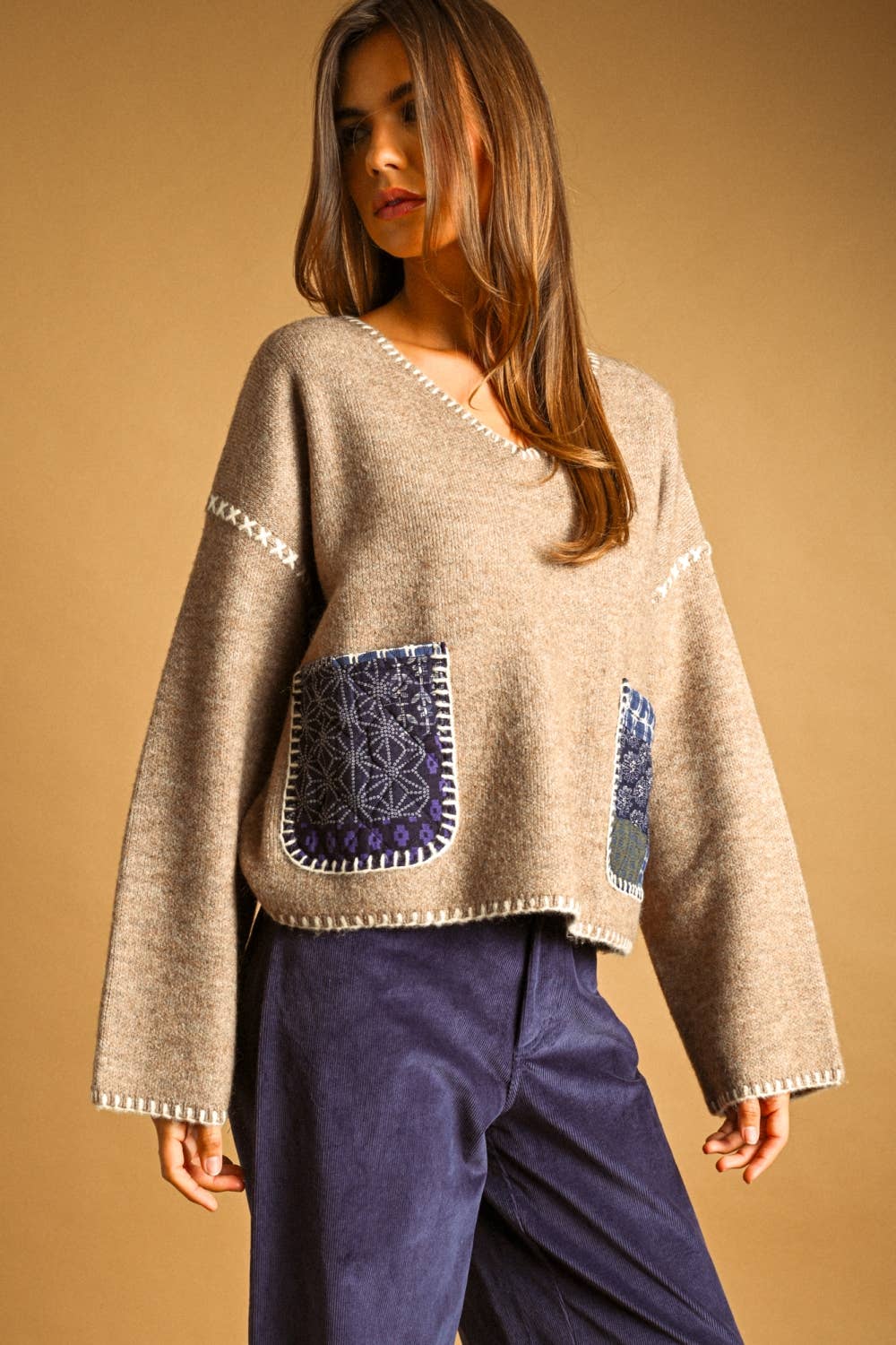 A woman wearing a beige knit sweater with decorative white trim and two dark blue patterned front pockets, paired with dark blue pants, against a solid tan background.