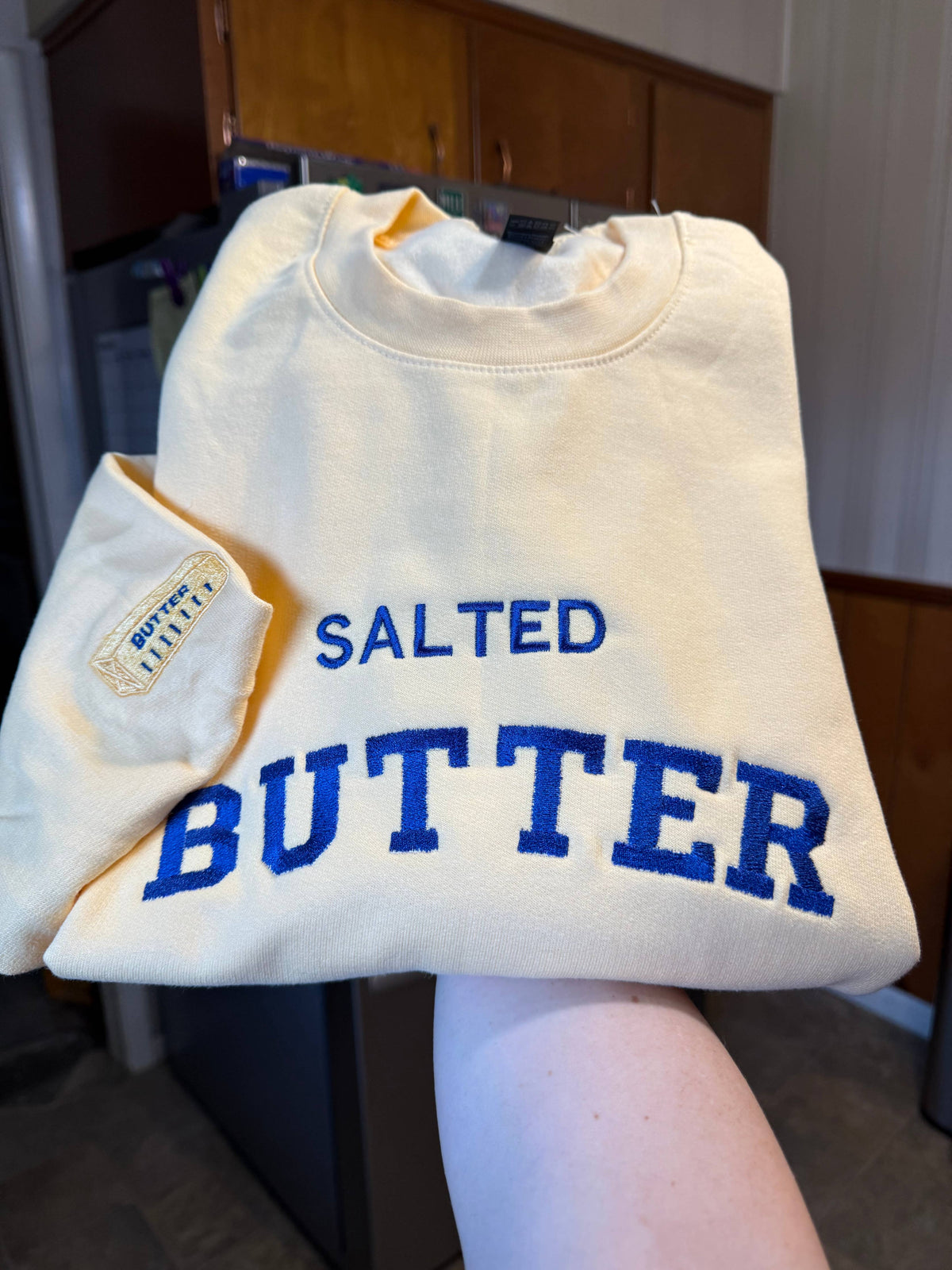 Yellow sweatshirt with 'Salted Butter' in blue text held by a person in a kitchen.