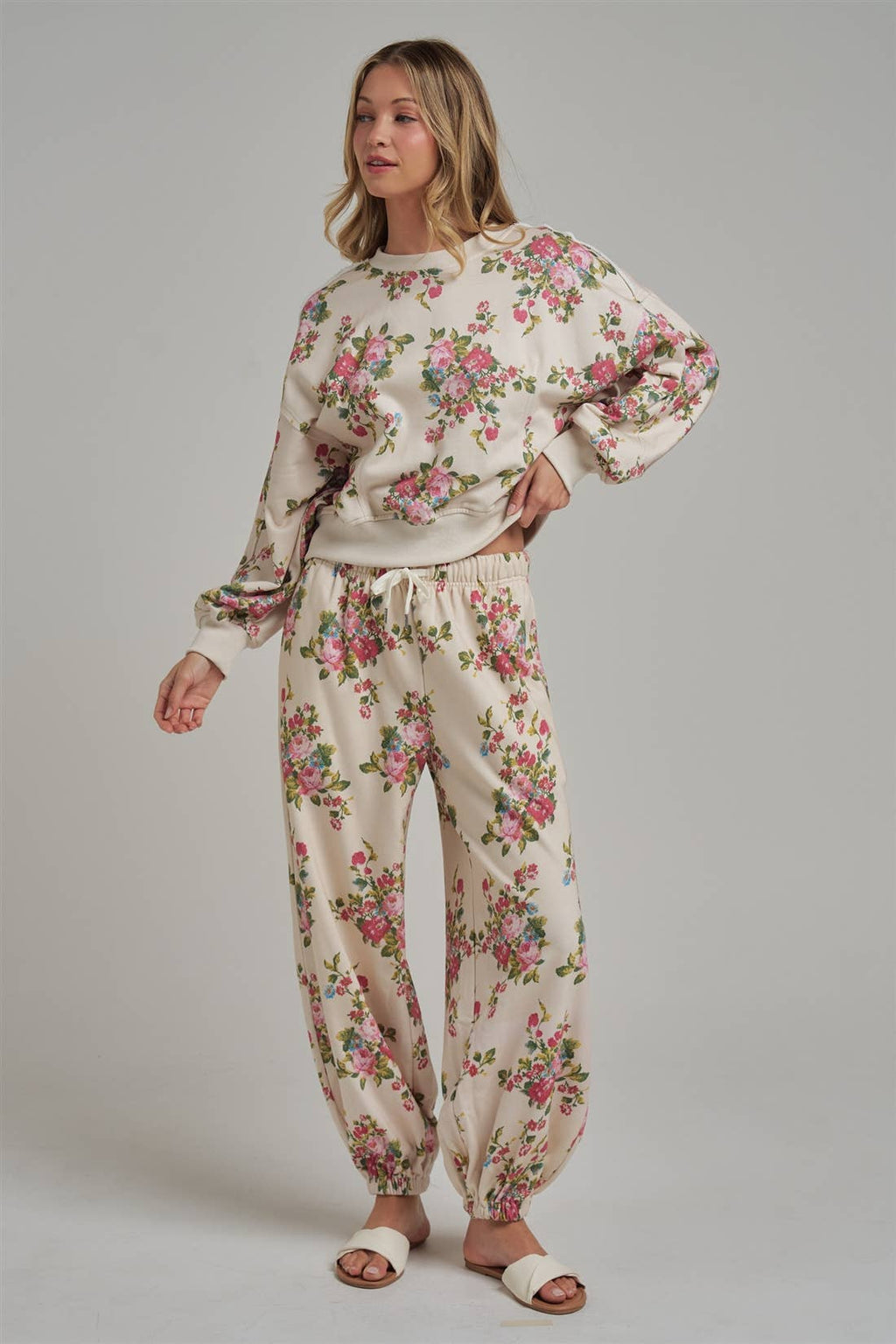 Floral patterned wide leg jogger sweat pants and matching pullover sweatshirt worn by a female model with white sandals on a plain background