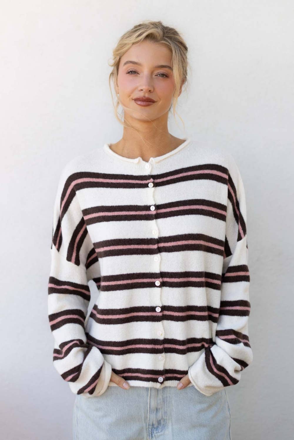 A woman wearing a cream-colored knit cardigan with horizontal dark brown and dusty pink stripes, paired with light-wash jeans, standing against a plain off-white wall.