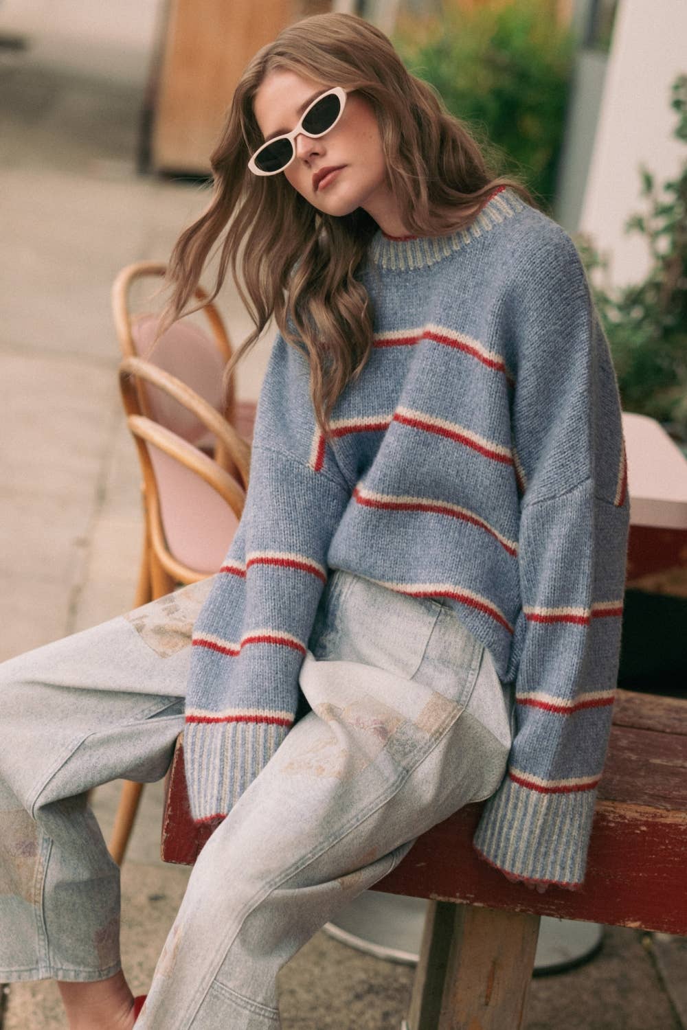 A woman wearing a light blue knit sweater with horizontal red and white stripes, paired with light-wash jeans and white-framed sunglasses, sitting on a wooden bench in an outdoor setting.
