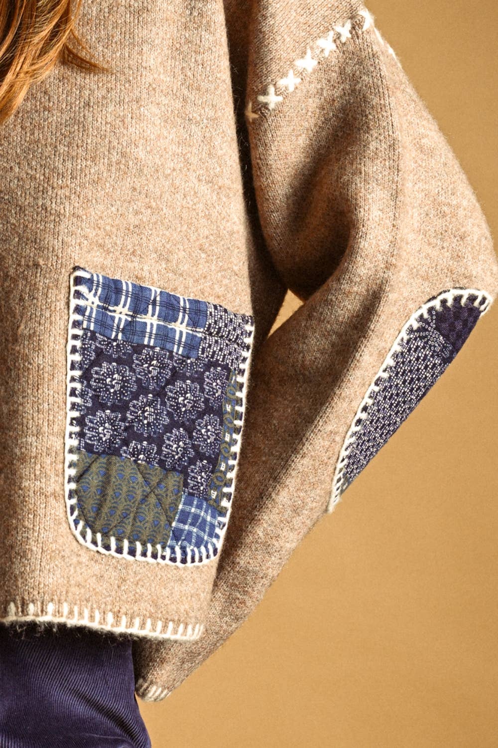 Close-up of a beige knit sweater with blue patchwork pockets and white contrast stitching, worn by a model against a warm tan background.
