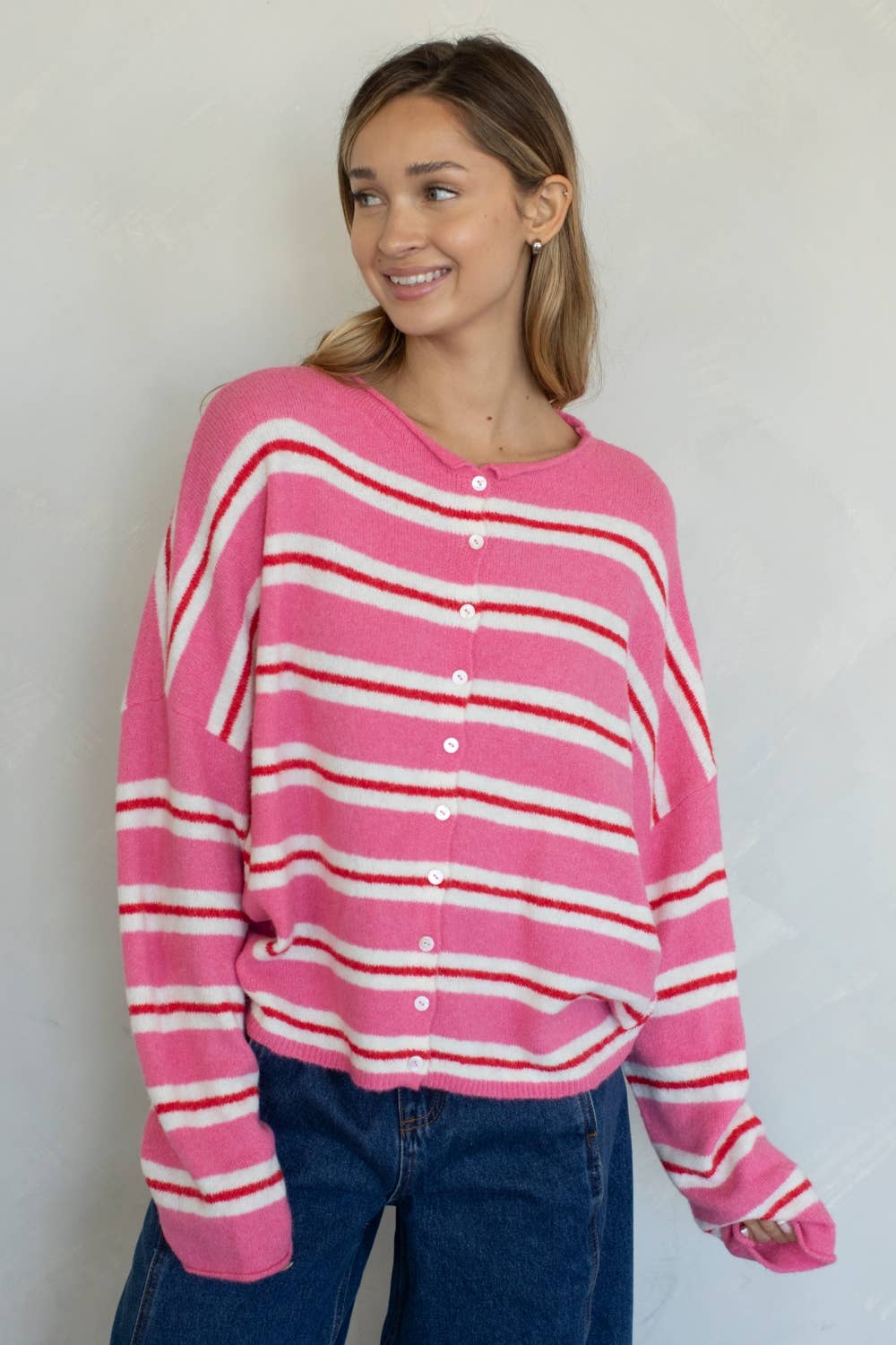 A woman wearing a bright pink knit cardigan with horizontal white and dark pink stripes, paired with darker denim jeans, standing against a plain off-white wall.