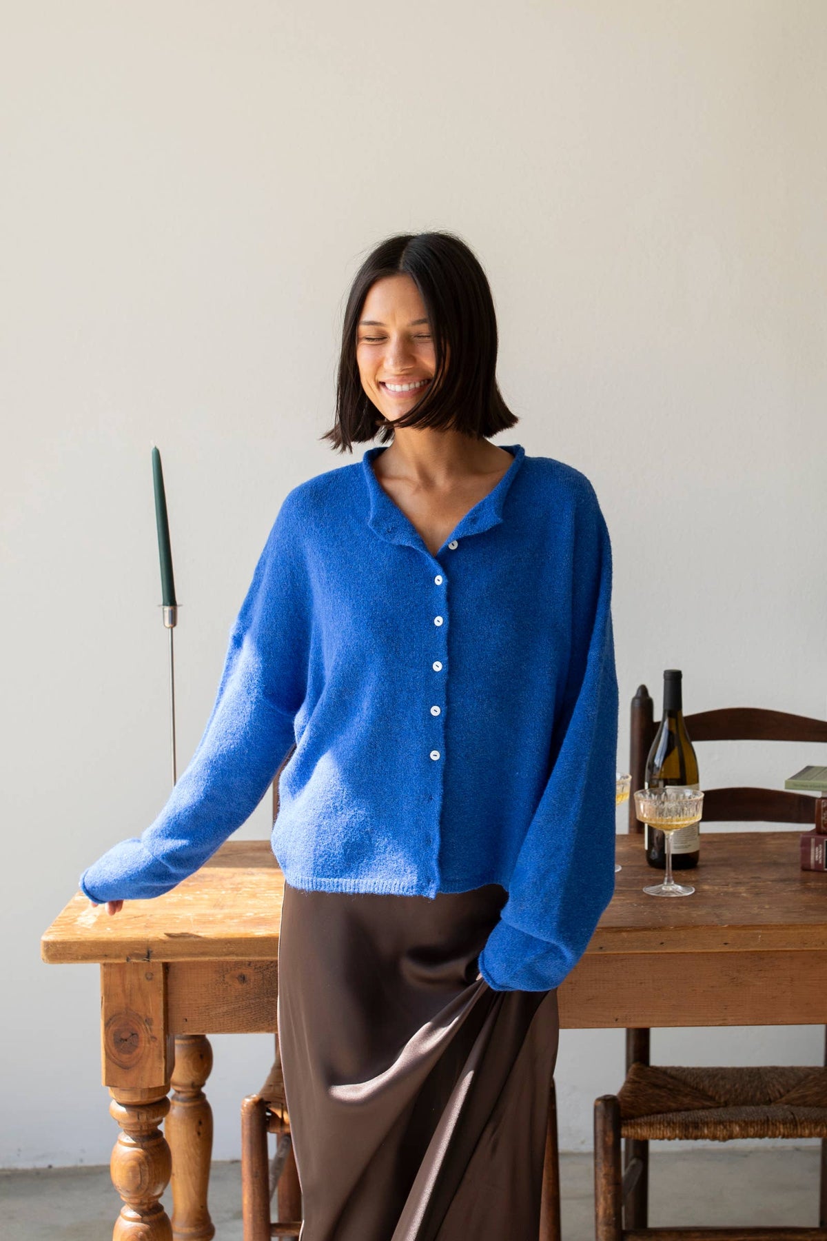 A female model wearing a bright blue button up cardigan with white buttons, paired with a brown silk skirt  in front of a table with chairs against a gray wall. 