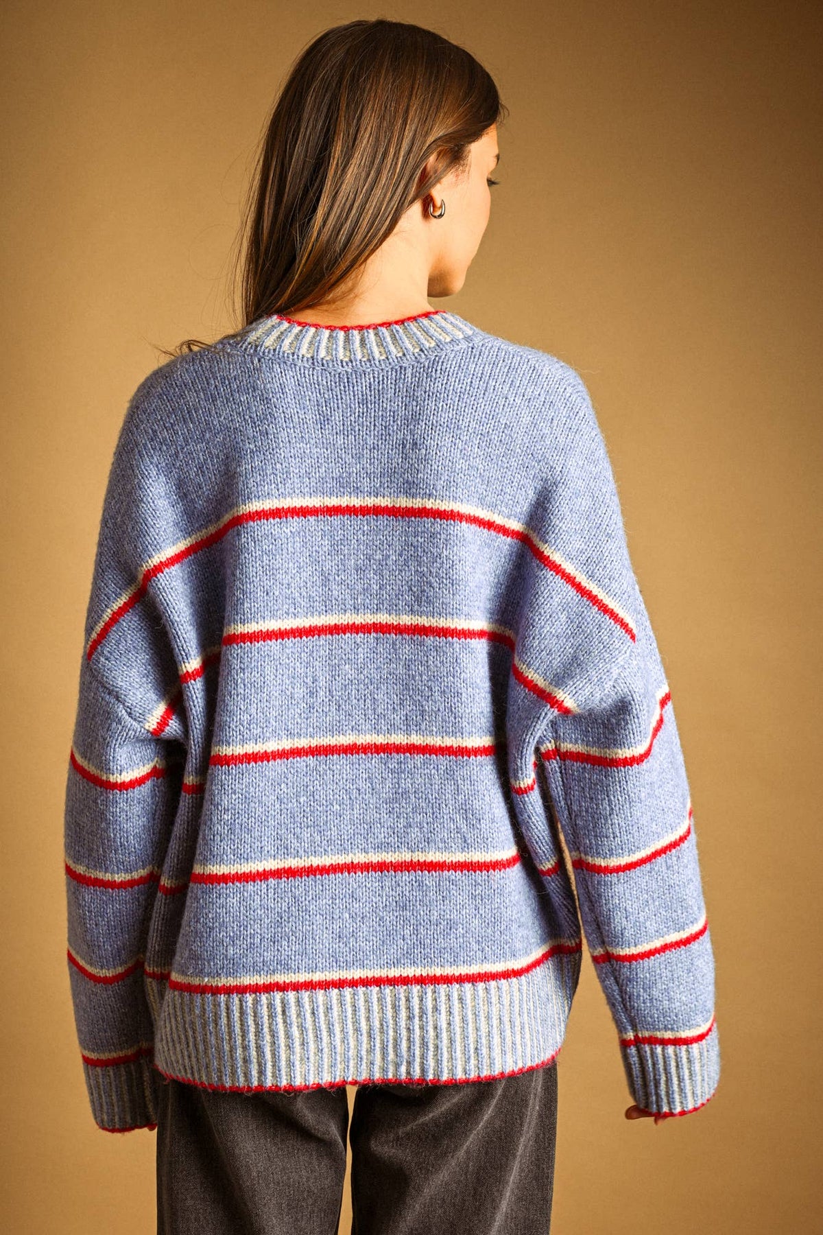 A woman viewed from behind wearing a light blue knit sweater with horizontal red and cream stripes, paired with dark gray pants, against a solid tan background.