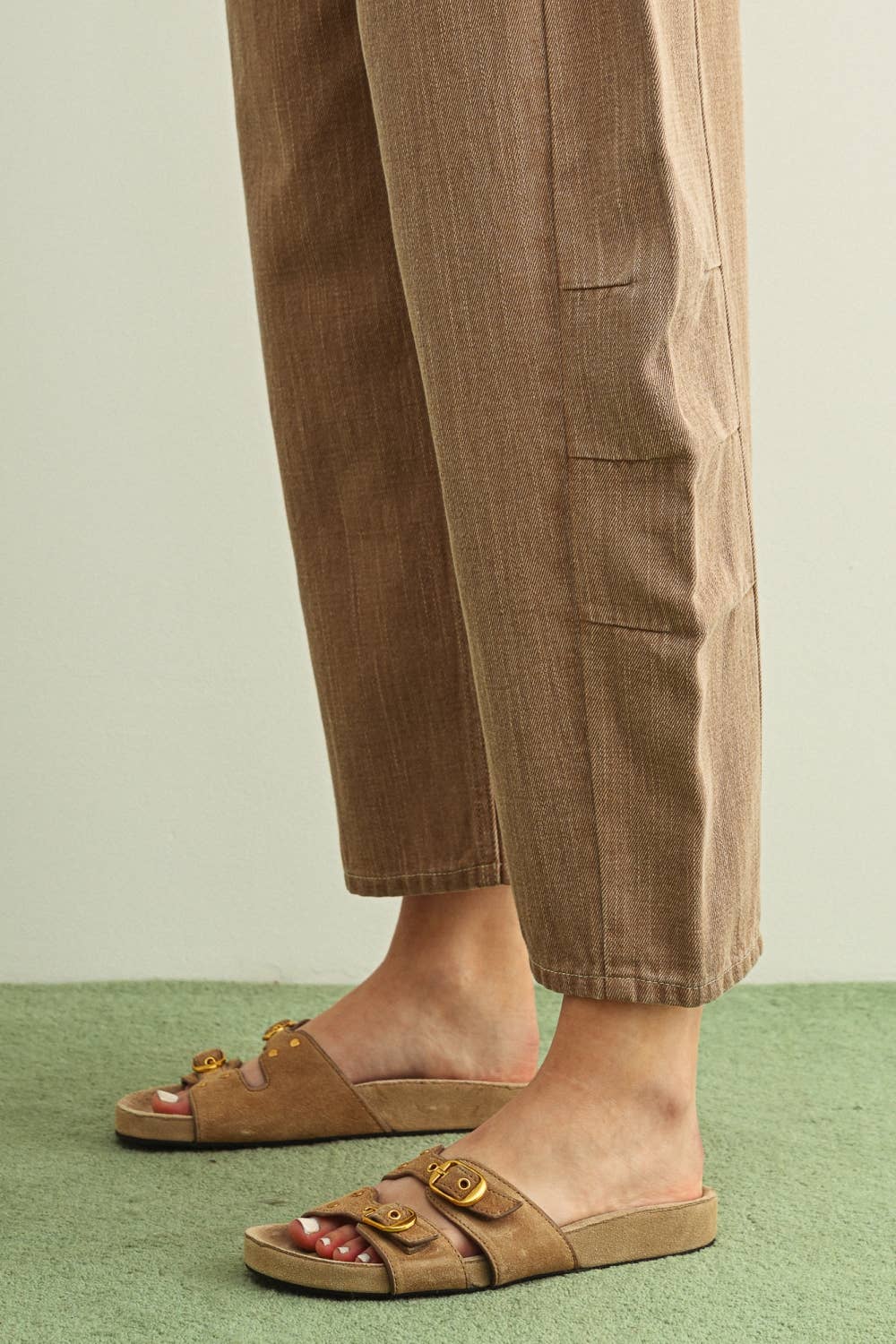 A close-up of a person's feet wearing brown double-strap slide sandals with gold-tone hardware, paired with matching brown barrel leg pants, standing on a green textured surface against a light beige wall.
