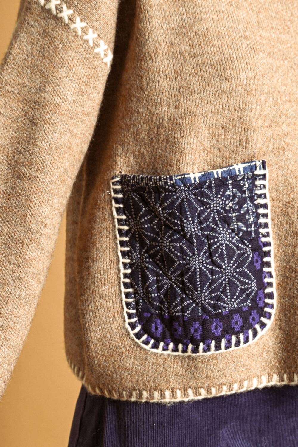 Close-up of a beige knit sweater with a decorative dark navy patch pocket featuring white geometric patterns and purple accents, shown on a model against a warm tan background.