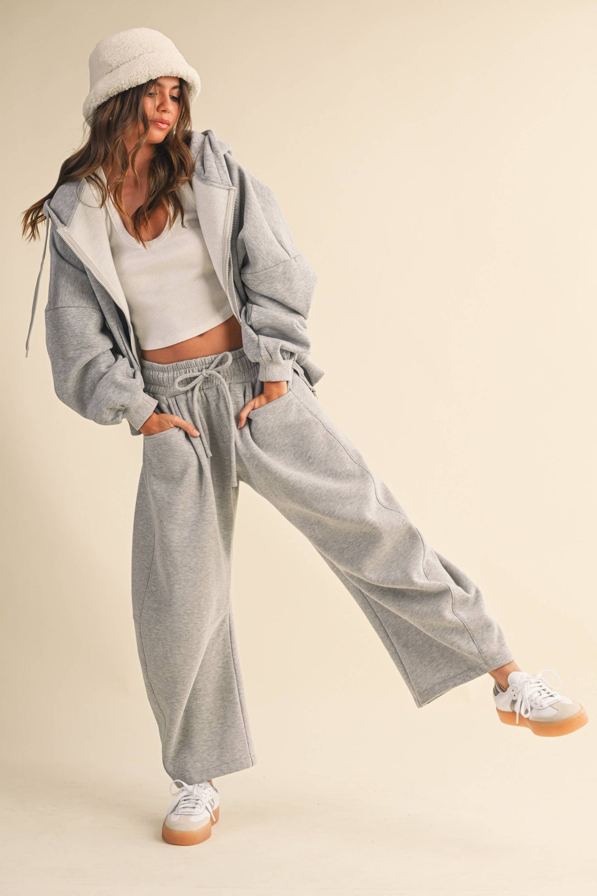 A woman modeling a light gray lounge set consisting of a zip-up hoodie and matching wide-leg barrel sweatpants, styled with a white crop top, white bucket hat, and white sneakers with tan soles, against a solid beige studio background.