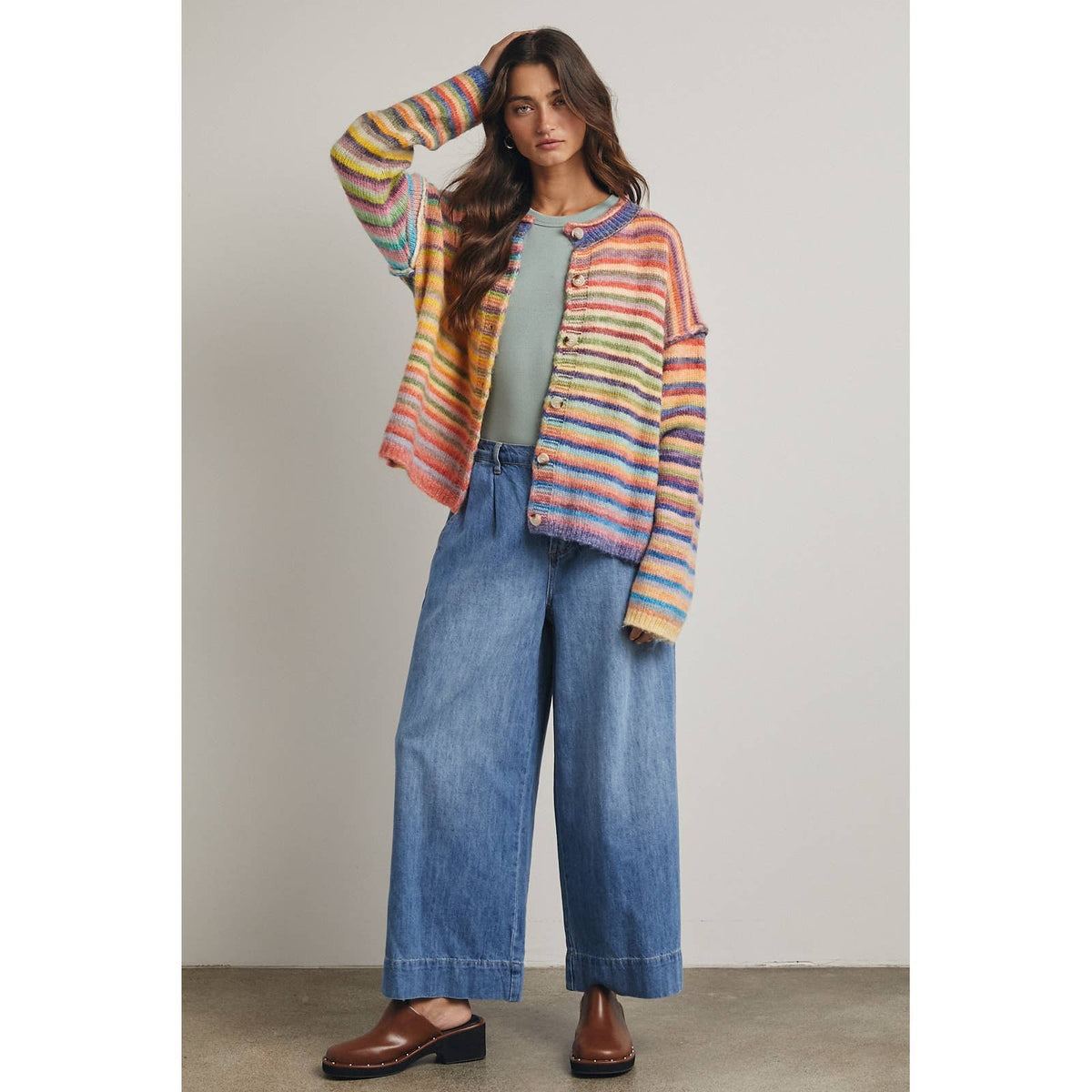 Woman wearing a colorful rainbow horizontally striped cardigan, green top, and blue jeans against a neutral background
