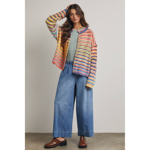 Woman wearing a colorful rainbow horizontally striped cardigan, green top, and blue jeans against a neutral background