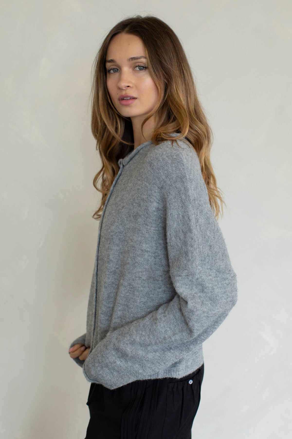 A female model facing sideways, wearing a light gray button up cardigan with white buttons, paired with black denim jeans in front of a gray studio background. 
