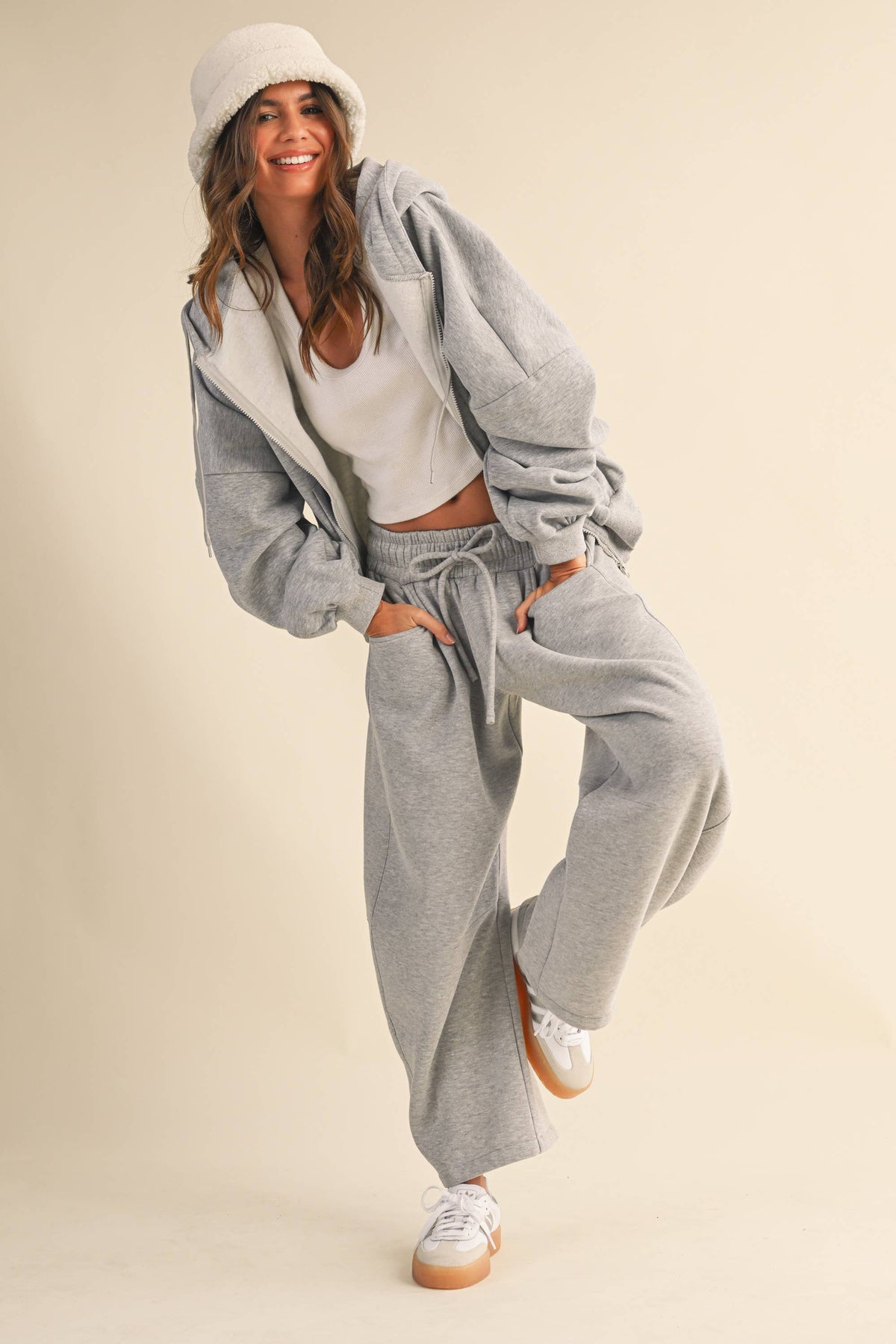 A woman modeling a light gray two-piece lounge set consisting of a zip-up hoodie and matching drawstring wide-leg barrel sweatpants, styled with a white crop top, white beanie, and white sneakers against a solid beige background.